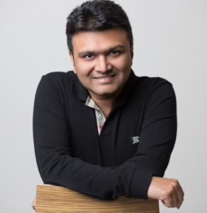 Smiling man in a black shirt leaning on a wooden surface against a plain white background.