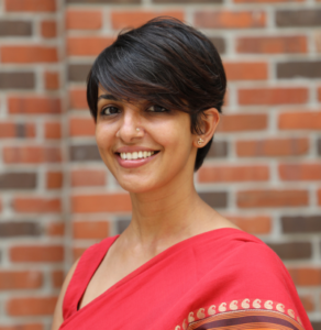 Smiling woman with short hair, wearing a red saree, standing in front of a brick wall.