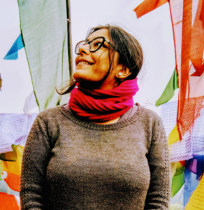 Smiling woman in glasses and red scarf, standing near colorful prayer flags in a bright outdoor setting.