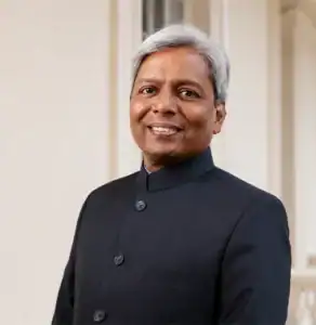 Smiling man with gray hair wearing a black suit, standing in front of a light-colored background.