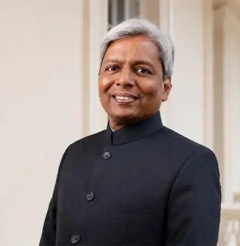Smiling man with gray hair wearing a black suit, standing in front of a light-colored background.