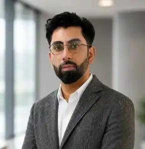 Man with glasses, beard, and gray suit jacket standing indoors near large windows.