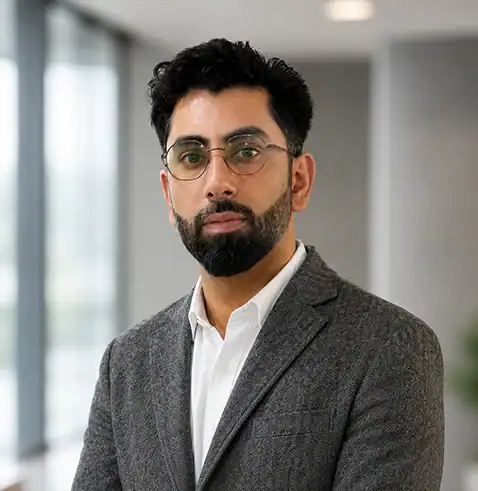 Man with glasses, beard, and gray suit jacket standing indoors near large windows.