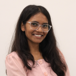 Smiling woman with long dark hair, clear glasses, pink blouse, and a white background.
