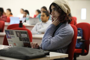 Person in a beige beanie focuses in a classroom, sitting at a laptop with stickers on it.