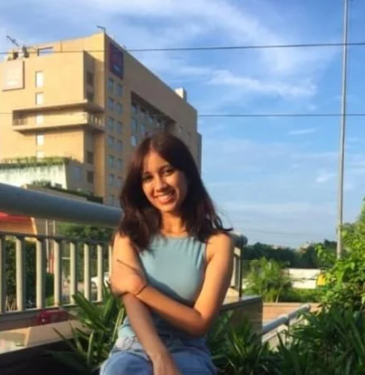 Smiling woman in a blue top sitting outdoors, surrounded by plants, with a building in the background.
