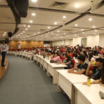 Speaker addressing a large audience seated in a well-lit auditorium with red chairs and white desks.