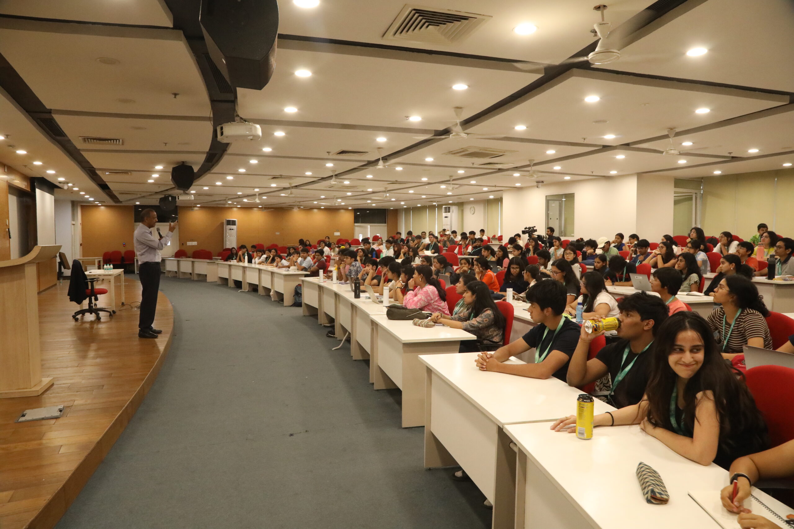 Speaker addressing a large audience seated in a well-lit auditorium with red chairs and white desks.