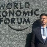 Man in a suit standing next to the World Economic Forum logo on a stone wall.