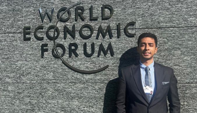Man in a suit standing next to the World Economic Forum logo on a stone wall.