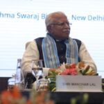 Man seated at a conference table with a nameplate reading "Shri Manohar Lal," floral centerpiece in front.
