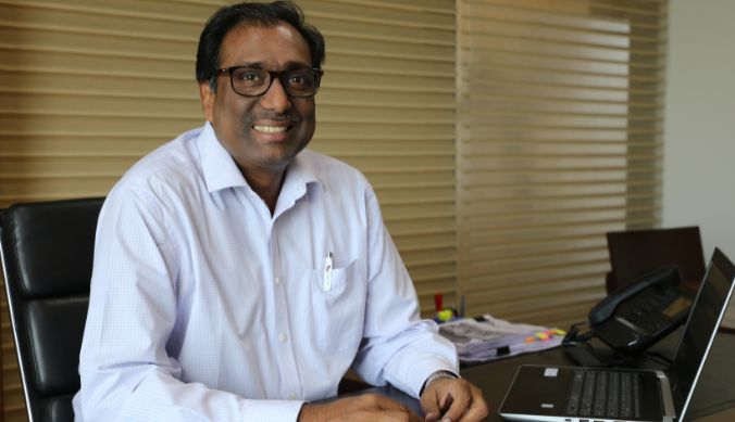 Smiling man in glasses and white shirt sitting at a desk with a laptop and office supplies.