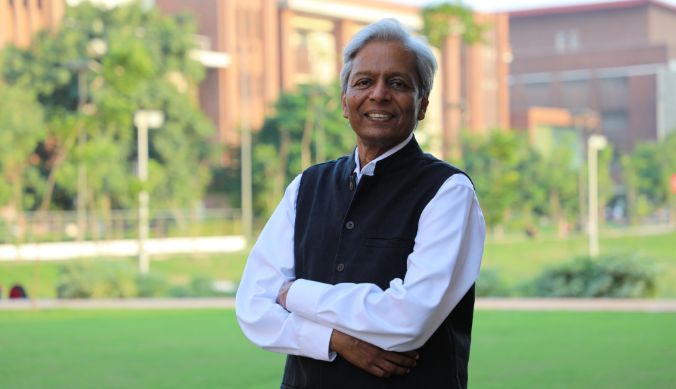 Smiling man in a black vest and white shirt stands outdoors with trees and buildings in the background.