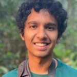 Smiling person with curly hair wearing a teal shirt and camera strap, greenery in the background.