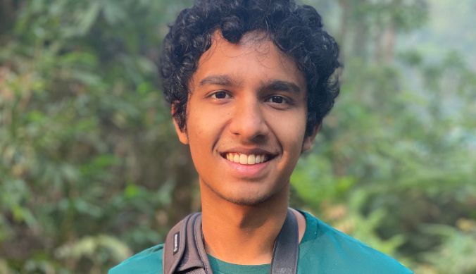 Smiling person with curly hair wearing a teal shirt and camera strap, greenery in the background.