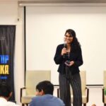 Woman speaking on stage with a microphone, banner reading "Startup Ashoka" on the left.