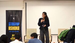 Woman speaking on stage with a microphone, banner reading "Startup Ashoka" on the left.