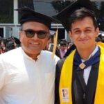Two men smiling at a graduation ceremony, one wearing a cap and gown with a yellow stole.