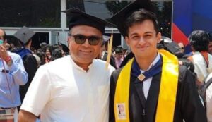 Two men smiling at a graduation ceremony, one wearing a cap and gown with a yellow stole.