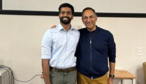 Two men standing together, smiling, one in a white shirt and the other in a navy sweater.