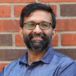 Smiling man with glasses and a beard, wearing a blue shirt, standing against a brick wall.