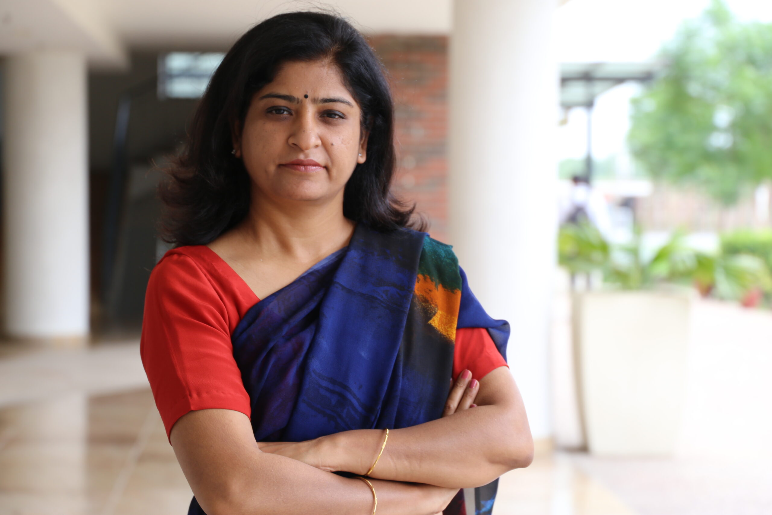 Woman in a blue saree with folded arms standing in an outdoor corridor with greenery in the background.