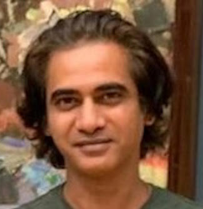 Man with wavy shoulder-length hair, wearing a green shirt, standing in front of a colorful background.