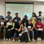 A group of 12 people sitting and standing on a stage, holding cards, smiling at the camera.