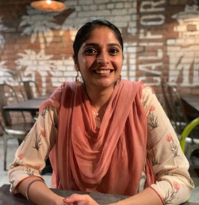 Smiling woman in a pink scarf seated at a table in a rustic café with brick walls and palm decor.