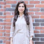 Woman in a beige tunic standing against a brick wall, smiling with long dark hair.