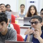 Students seated in a classroom, one holding a microphone, with laptops on desks.