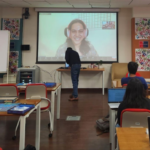 Classroom with students using laptops, instructor at the front, large screen showing a smiling person.