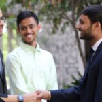 People in formal attire interacting outdoors, two shaking hands, others smiling in the background.