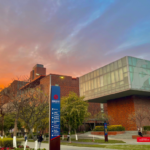 Modern building with glass and brick facade, trees, pathway, and vibrant orange-blue sunset sky.