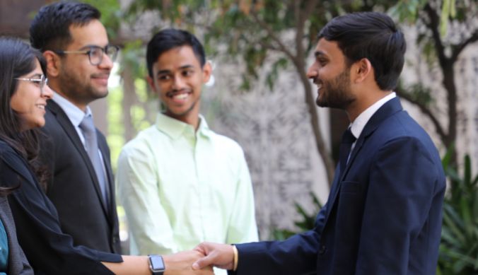 People in formal attire interacting outdoors, two shaking hands, others smiling in the background.