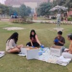 Four people sorting items from boxes on a grassy lawn with buildings and trees in the background.