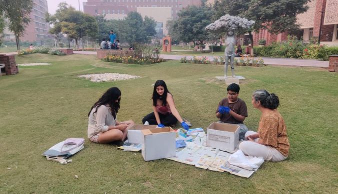 Four people sorting items from boxes on a grassy lawn with buildings and trees in the background.