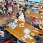 Students playing cards around a wooden table in a classroom, with others seated in the background.