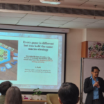 Speaker presenting a slide about strategy with a board game illustration to an audience in a classroom.