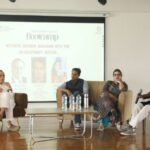 Panel discussion with four speakers seated on a stage, projector screen in the background.