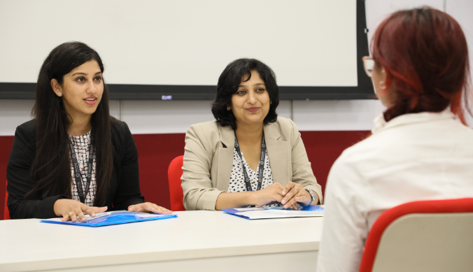 Two women conducting an interview at a table, facing a candidate with red hair in a white shirt.