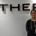 Woman in black embroidered top standing near a wall with "ATHER" logo in bold black letters.