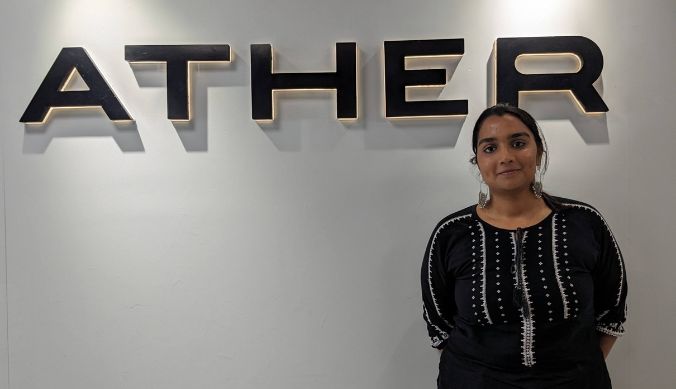 Woman in black embroidered top standing near a wall with "ATHER" logo in bold black letters.