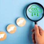 Hand holding magnifying glass over CO2 cloud icon, three wooden circles with white arrows below.