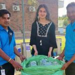 Three people holding a green bag filled with plastic bottles outdoors near recycling bins.