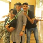 Three smiling people posing playfully indoors, one making a peace sign, with a seated person nearby.