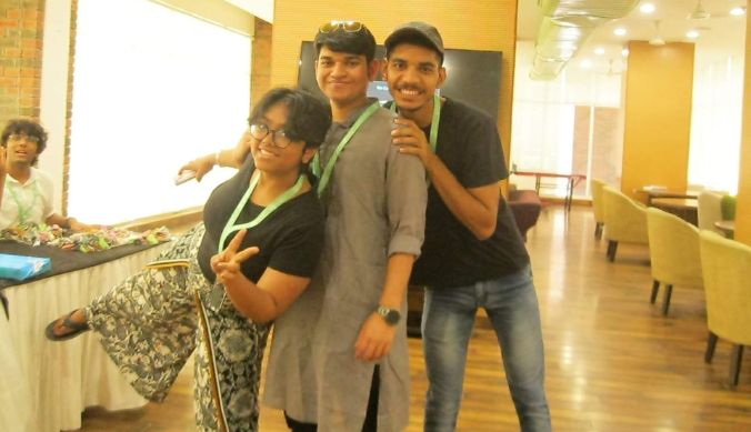 Three smiling people posing playfully indoors, one making a peace sign, with a seated person nearby.