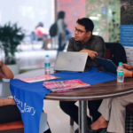 Three people seated at a table with laptops and brochures, engaged in a discussion indoors.