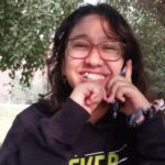 Smiling person with glasses and dark hair, holding a phone outdoors near trees and seating.