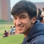 Smiling person in a blue hoodie sitting on grass, with people and buildings in the background.